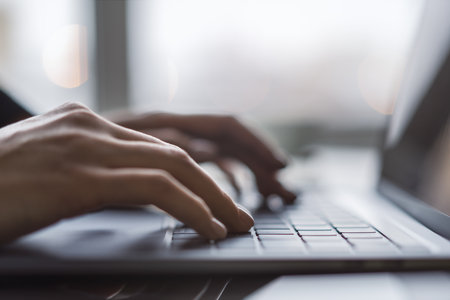 Against a blurred office background, the image captures a woman's hands typing on a sleek laptop keyboardの写真素材