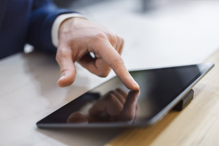 Man's finger engaging with a cutting-edge digital tablet screen, device situated on a desk within the blurred contours of a sunlit officeの写真素材