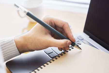 Close-up depiction of a woman's hand taking notes in a notepad on a sleek laptop, with a blurred...