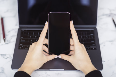 Close detail of a woman operating her latest-model smartphone, laptop lying open adjacent surrounded...