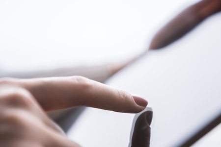 Closeup view of a female finger operating a high-tech tablet on a desk, set against the blurred backdrop of a luminous office sceneの写真素材