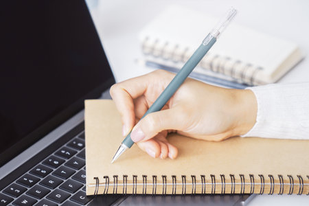 Close-up capture of a woman's hand as she writes in a notepad on a sleek laptop, with an...