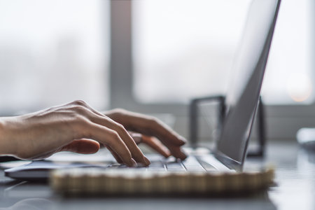 In a blurry office environment, a close-up view reveals a woman's hands typing on a trendy laptop...