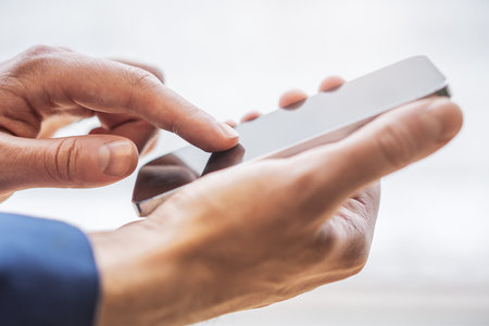Close detail of a man operating his latest-model smartphone surrounded by the unfocused hustle of his officeの写真素材