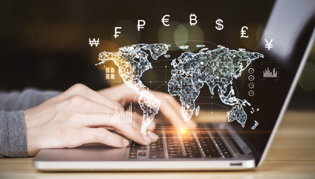 Hands typing on laptop with holographic world map and currency symbols overlay, glowing digital...
