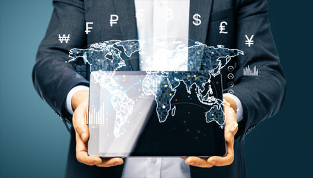 Businessman holding tablet displaying holographic world map with currency symbols and digital data on blue background, concept of financeの写真素材