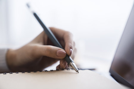 A woman's hand is caught in close-up, busily writing in a notepad on a laptop, while the background is out of focusの写真素材