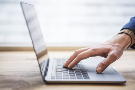 The picture shows a close-up of a man's hands typing on a contemporary laptop keyboard, with a softly blurred office setting in the backdropの写真素材