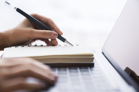 In close view, a woman's hand writing down notes in a notepad on a laptop, against a hazy backdropの写真素材