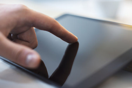 Focused view of a male hand interacting with a modern tablet device on a desk, amid the indistinct...
