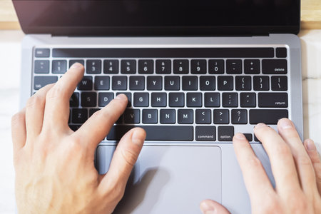 A close-up reveals a man's hands swiftly typing on the keyboard of a modern laptop, with a blurred office setting behindの写真素材