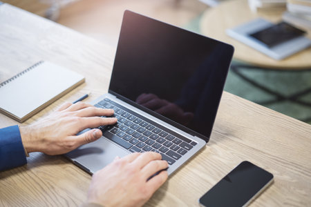 In a close-up shot, a man's hands are seen deftly typing on a modern laptop keyboard, set against a blurry office backdropの写真素材