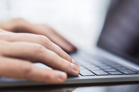 A man's hands can be seen in close detail, typing on a trendy laptop keyboard, with a blurry office...