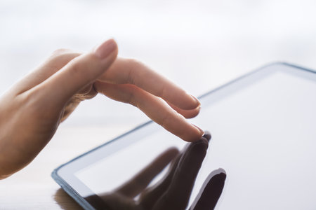 Detailed observation of a woman's finger navigating the screen of an advanced tablet, device placed on a desk against the vague outline of a well-lit officeの写真素材