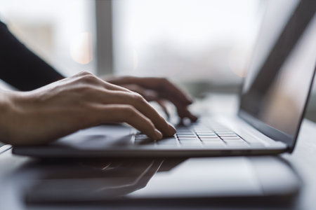 Against a blurred office background, the image captures a woman's hands typing on a sleek laptop keyboardの写真素材