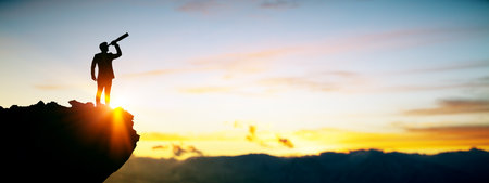 Silhouette of business person with telescope on mountain top at sunrise, with warm light and sky background, concept of vision and leadershipの写真素材