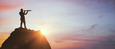Silhouette of a business person using a telescope atop a mountain, natural sunrise background, concept of vision, leadership, and goal settingの写真素材