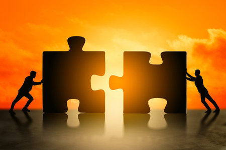Silhouettes of two people pushing large puzzle pieces together on glowing orange background, symbolizing teamwork, connection, and collaborationの写真素材