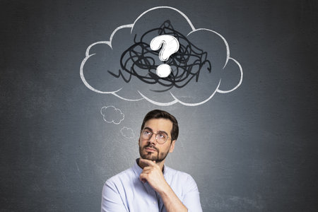 Businessman thinking with question mark in cloud symbol above head, hand on chin, on dark wall background, concept of brainstorming or problemの写真素材
