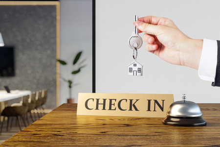 Closeup of a person holding a house key above a reception counter with a check-in sign and bell, in a modern hotel interior background conceptの写真素材