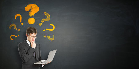 Thoughtful person in suit holding laptop, yellow question marks above head, dark chalkboard background, concept of confusion or decision-makingの写真素材