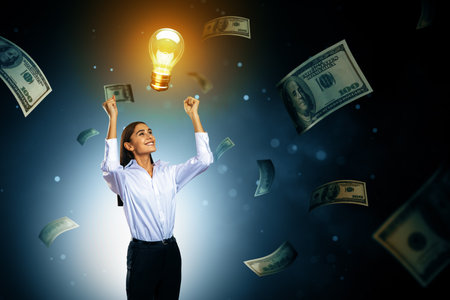 Confident businesswoman celebrates bright idea with glowing bulb and flying money on dark background, symbolizing success and financial innovationの写真素材