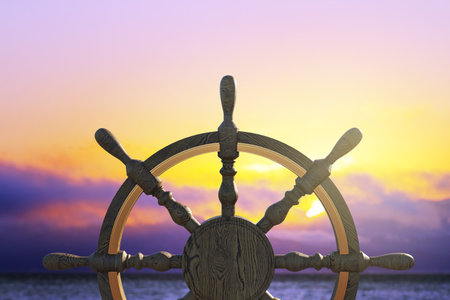 Wooden ship wheel in close-up with ocean and dramatic sunset sky background, concept of travel, journey, vacation, and nautical explorationの写真素材