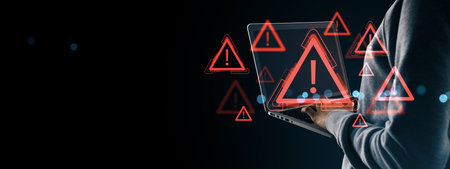 Person in hoodie using laptop with red warning signs, exclamation marks and digital alerts on dark background, symbolizing online fraud or dangerの写真素材