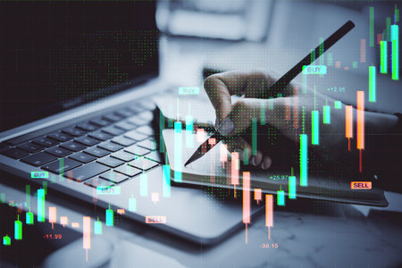 Hand writing on notepad beside laptop with digital forex candlestick chart overlay on office desk, concept of financial market analysisの写真素材