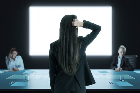 Businesswoman standing with hand on head in dark office conference room, back view, facing bright screen during corporate meeting conceptの写真素材