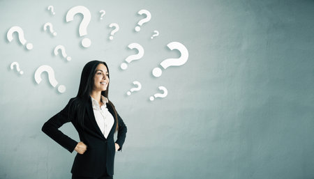 Smiling businesswoman in formal suit with question mark symbols around her on a light wall...