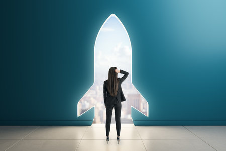 Woman in business suit stands before rocket-shaped door cutout in wall, looking out to city skyline, symbolizing startup growth and ambitionの写真素材