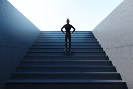 Female businessperson viewed from behind standing at the bottom of stairs in concrete space, symbolizing ambition, career and future successの写真素材