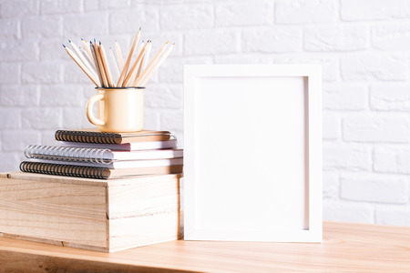 Desk with blank white picture frame and pencils in iron mug placed on books and wooden box. Mock up