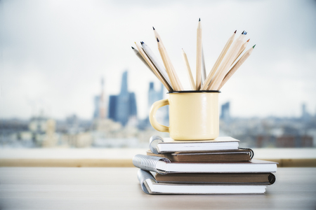 Iron mug with wooden pencils on stack of copybooks and blurry city in the backgroundの写真素材