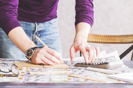 Male hand using calculator and writing in notepad on desktop covered with dollar banknotesの写真素材