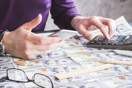 Closeup of man using phone and calculator on wooden table covered with dollar banknotesの写真素材