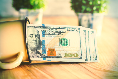Lying iron mug with dollar stack on wooden table with blurry plants in the backgroundの写真素材