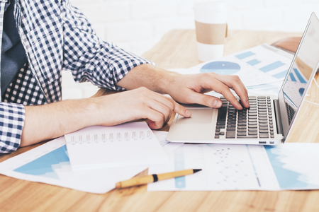 Sideview of businessman using laptop on desk full of paper sheets with business reports and coffee cupの写真素材