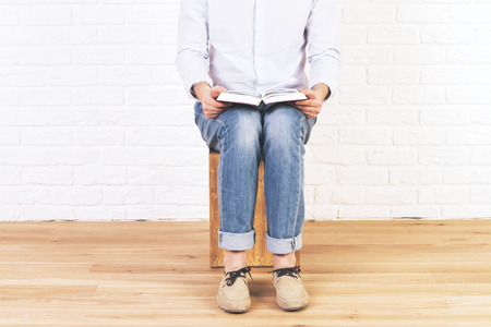 Caucasian male sitting in empty brick interior with wooden floor, reading book placed on his lapの写真素材