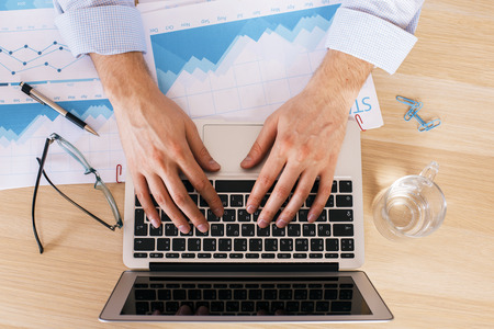 Male hands typing on keyboard of laptop placed on wooden desk with business reportの写真素材