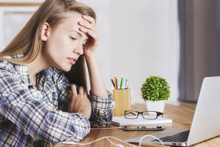 Closeup of tired caucasian female sitting at wooden office desk looking at computer screenの写真素材