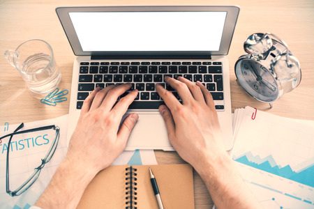 Topview of male hands typing on keyboard of laptop with blank white screen on office desk with glass of water and alarm. Toned image, Mock upの写真素材