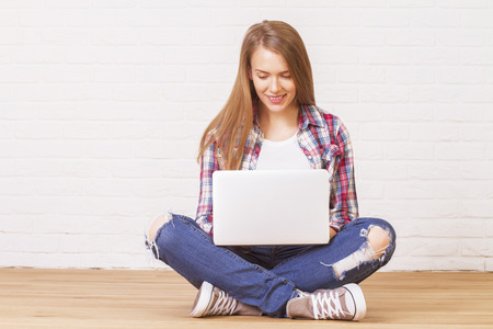 Smiling caucasian female sitting on wooden floor in studio and using laptopの写真素材
