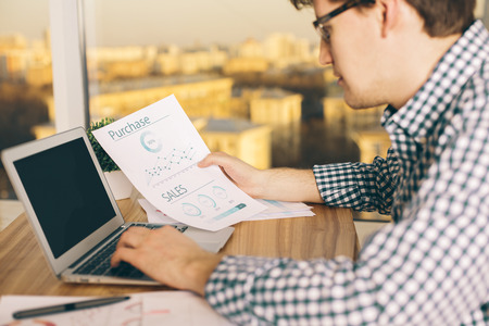Closeup of young caucasian male copying information from business report to his computer with blank screen. Sunlit city background. Mock upの写真素材