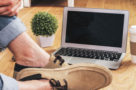 Male feet on wooden floor next to blank notebook screen, coffee, plant and other items. Mock upの写真素材