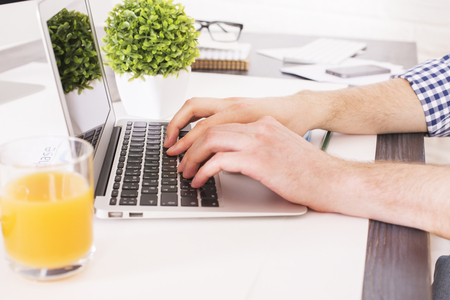 Sideview of businessman hands typing on laptop keyboard placed on office desktop with orange juice, plant and other itemsの写真素材