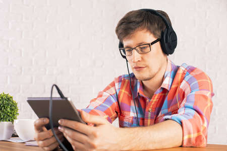 Caucasian male sitting at wooden desk and listening to music on his tablet. Mock up