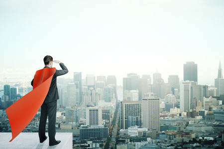 Businessman with red superhero cape standing on pedestal and looking into the distance on cityscape backgroundの写真素材