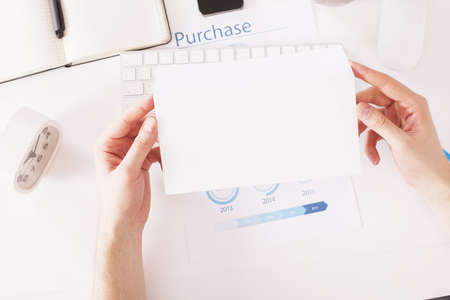 Hands holding regular blank envelope over office desktop with keyboard, clock, business report and other items. Topview, Mock upの写真素材